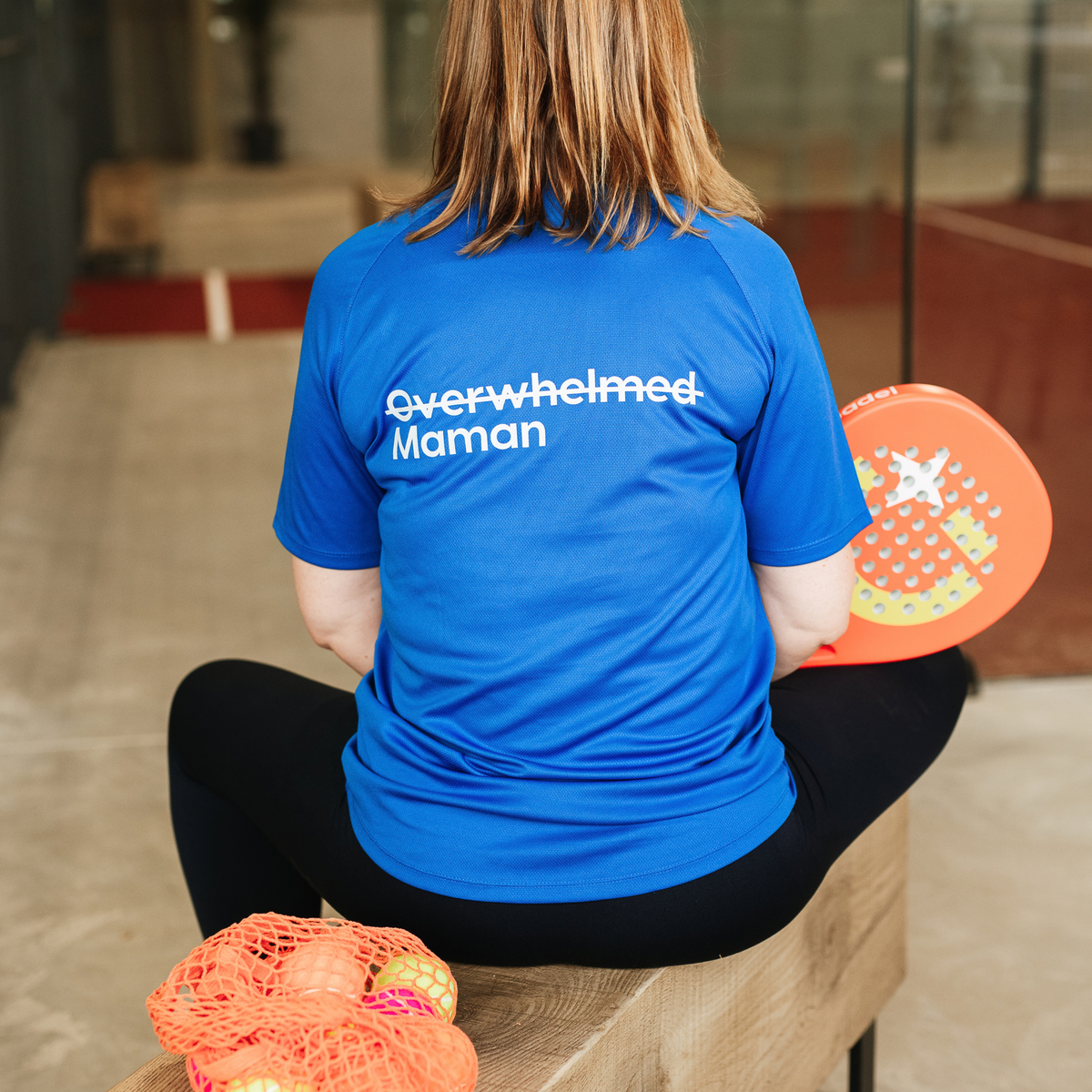 A woman sitting on a bench from behind, wearing a bright blue t-shirt with the text 'Overwhelmed Maman' printed in white on the back. 
