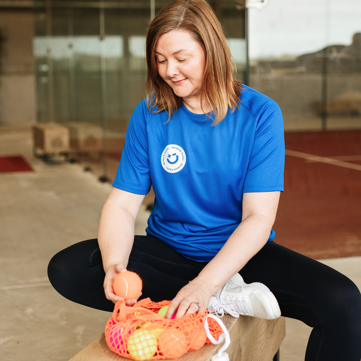 A woman in a bright blue t-shirt with a circular logo sits on a bench at an indoor court, holding an orange mesh bag filled with balls.