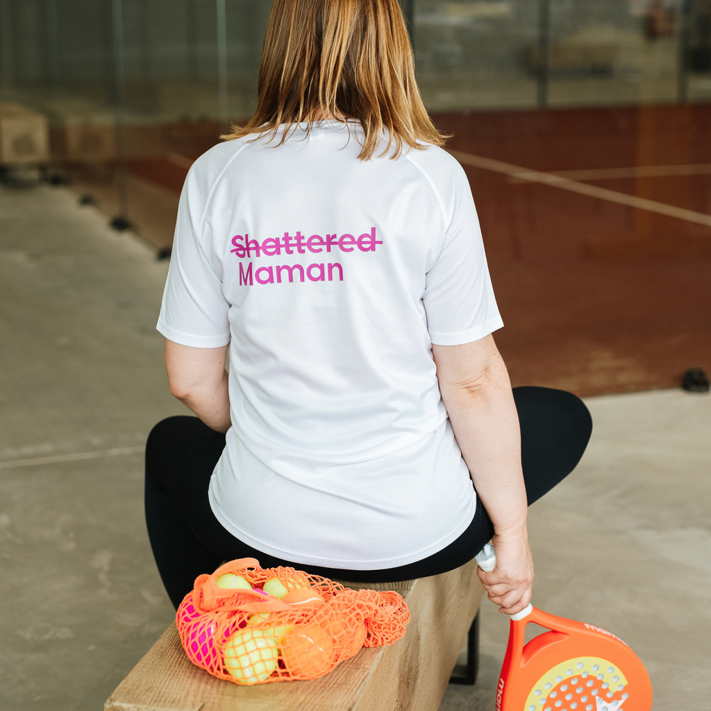 A woman sits on a wooden bench in an indoor sports court, viewed from behind. She is wearing a white short-sleeve athletic shirt with 'Shattered Maman' printed in pink across the back, paired with black leggings.