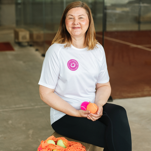 A woman sitting on a bench in an indoor gym or court space, wearing a white t-shirt with a pink circular logo on the left chest. She is holding two balls, one pink and one orange, and has a mesh bag of  balls in front of her.