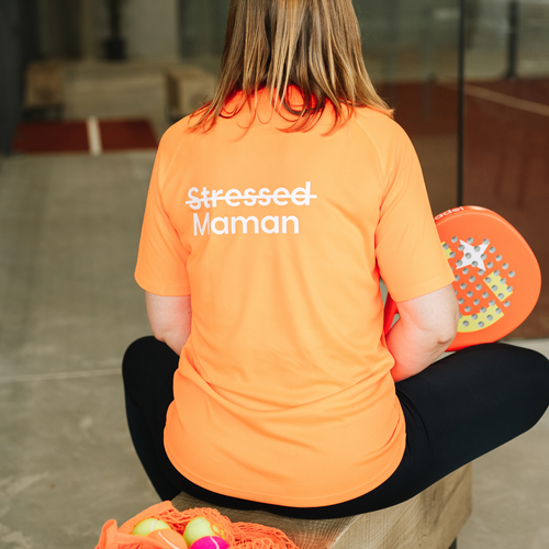 A woman sitting with her back to the camera wearing a bright orange t-shirt with 'Stressed Maman' printed in white on the back, holding a colorful padel paddle.