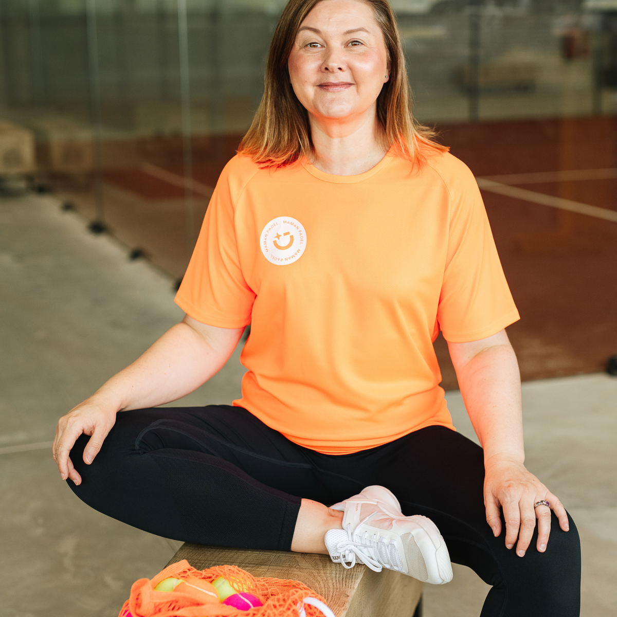 A woman sitting cross-legged in a gym wearing a bright orange athletic t-shirt with a white circular logo on the left chest, paired with black leggings and white sneakers.